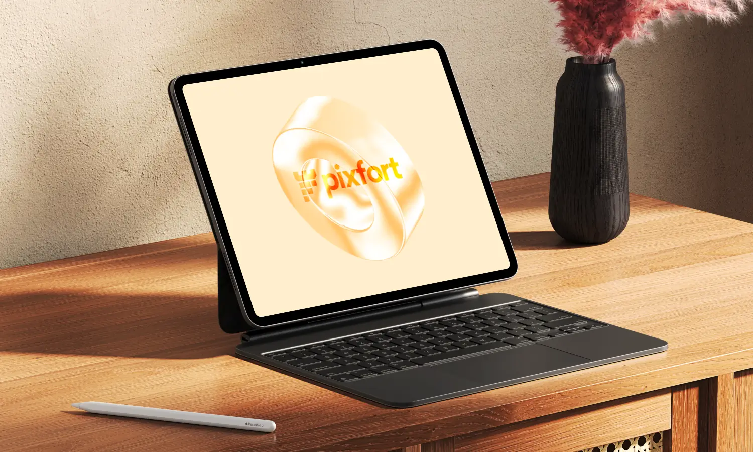 Tablet with detachable keyboard on a wooden desk, beige screen showing a logo, a white stylus nearby, and a dark vase with pink flowers to the right.