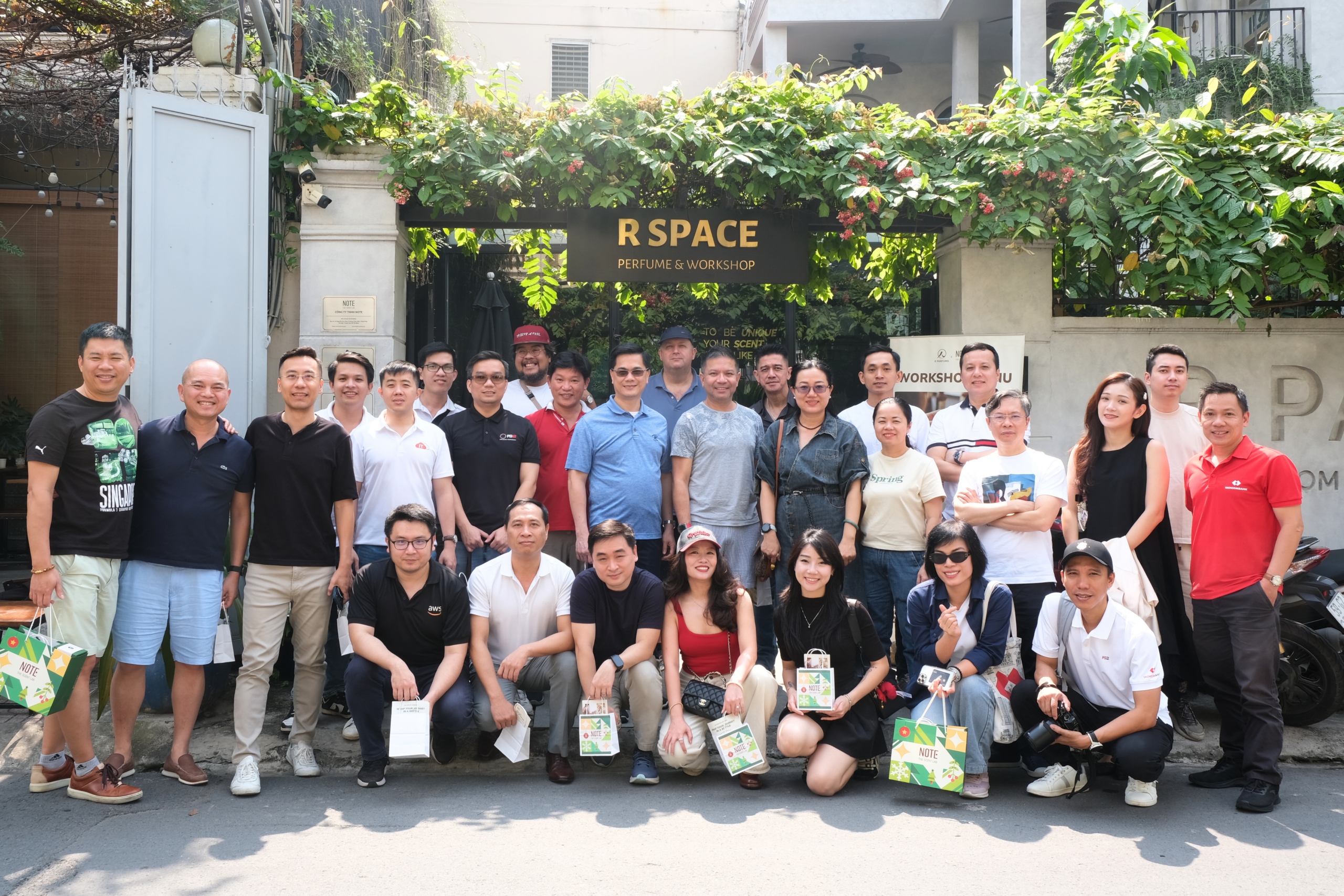 Group of people posing outdoors in front of a shop sign that reads 'R SPACE Perfume & Workshop' with green vines overhead.