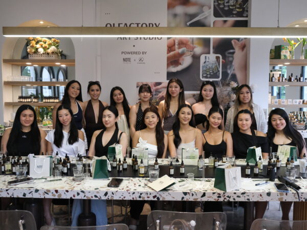 Group of about 15 women posing together behind a makeup table at a beauty event, with products and gift bags on the counter and a promotional backdrop behind them.