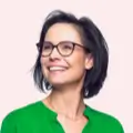 Smiling woman with short dark hair and glasses wearing a green blouse against a pale pink background.