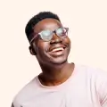 Smiling man with short hair and clear-framed glasses, wearing a light pink shirt against a pale beige background.