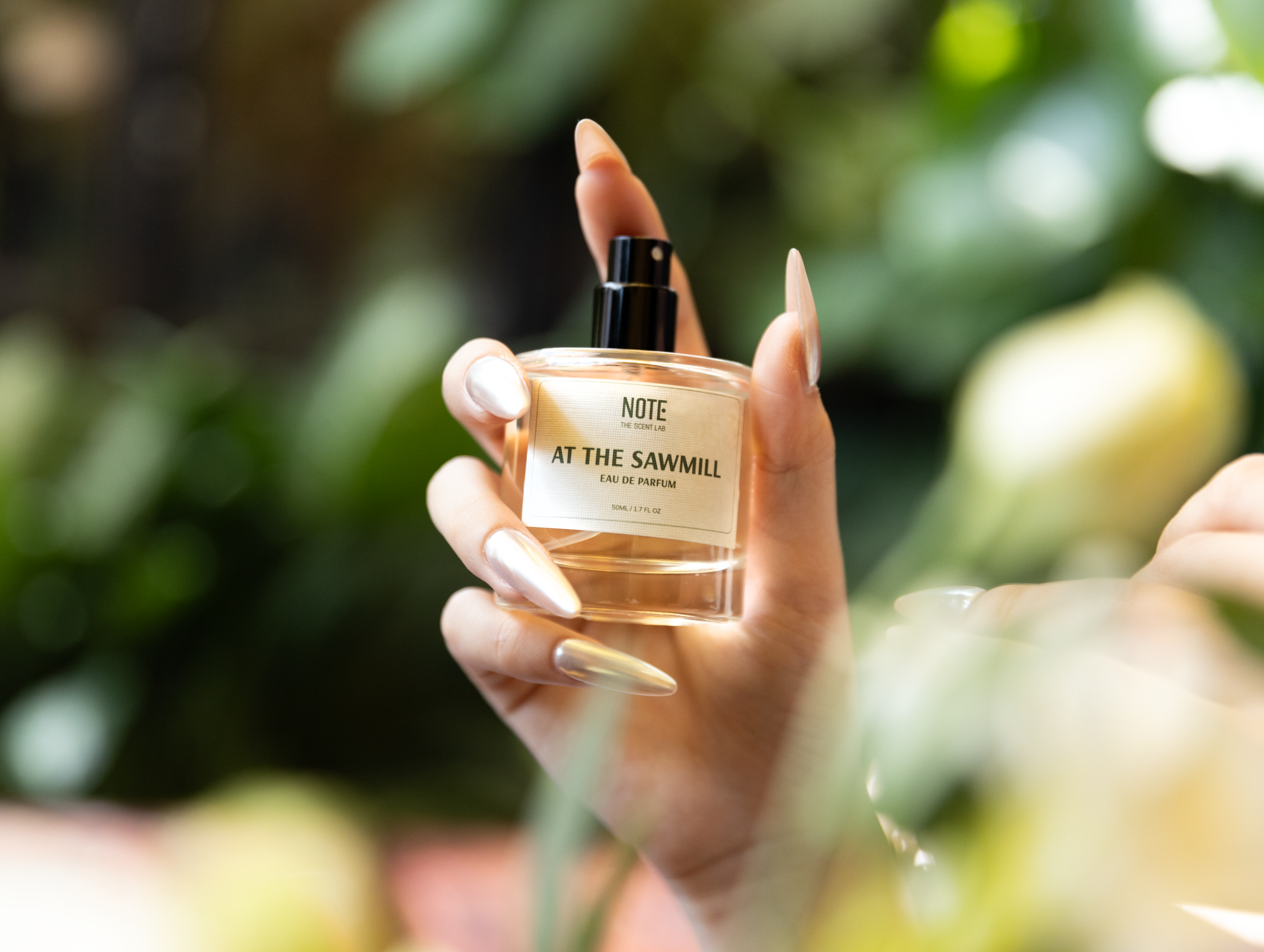 Small perfume bottle labeled 'AT THE SAWMILL' being held between two manicured fingers against a blurred green background (outdoors).
